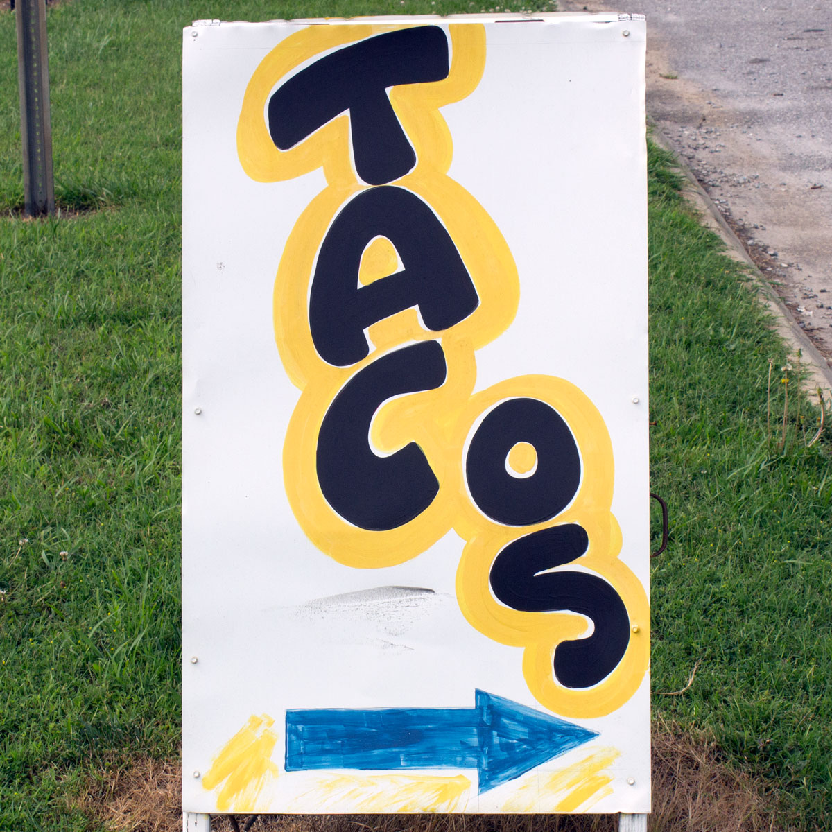 Sandwich board sign that reads ‘tacos’ spelled out vertically but with ‘o’ and ‘s’ placed to the right of ‘t’, ‘a’, and ‘c’